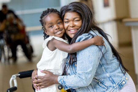 A Black pediatric patient hugs her mom. The patient wears braces on her legs and has a child-sized walker.
